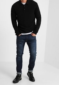 Svart långärmad pikétröja med krage, i kombination med mörkblå slim fit jeans och svarta sneakers med texturerad yta.