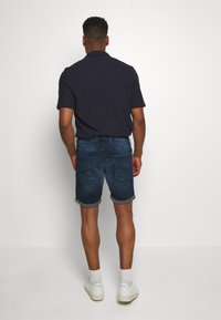 Tummansiniset farkkushortsit, käännetyt lahkeet, kahdella takataskulla, yhdistettyinä tummansiniseen polo-paidan kanssa. Valkoiset lenkkarit ja ribbisukat näkyvillä.