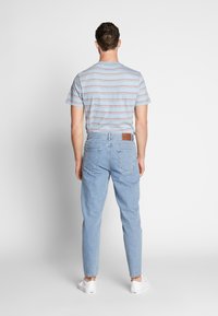 Młody mężczyzna w paskowanej koszulce z krótkim rękawem, jasnoniebieskich dżinsach i białych trampkach, stojący na tle jednolitego białego tła.