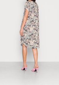 Mujer que lleva un vestido suelto de manga corta con un patrón de paisley en tonos pastel, combinado con sandalias de tacón abierto rosas, de pie sobre un suelo gris.