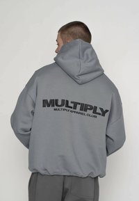 Šedá mikina s kapucňou a voľným strihom, s výrazným čiernym logom „MULTIPLY APPAREL CLUB“ na chrbte a elastickým lemom.
