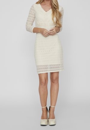 Kobieta ubrana w kremową koronkową sukienkę do kolan z rękawami do połowy przedramienia oraz kremowe platformy na obcasie z paskami wokół kostek.