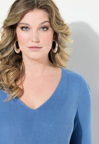 Blaues geripptes Strickoberteil mit V-Ausschnitt, langlebigen Ärmeln und strukturierter Oberfläche. Das Model trägt große Creolen. Der Hintergrund ist schlicht.