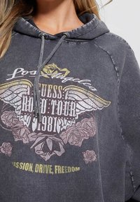 Femme portant un sweat à capuche gris foncé avec l'inscription "Los Angeles", des ailes, des roses, et les textes "Guess Road Tour 1981" et "Passion, Drive, Freedom".