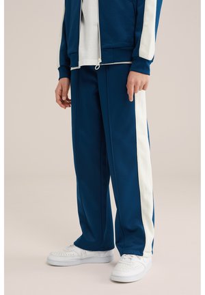 Person iført marineblå træningsbukser med hvide sidelommer og hvide sneakers, kombineret med en matchende jakke og hvid skjorte.