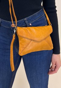 Borsa a tracolla in pelle arancione con patta pieghevole, superficie testurizzata e tracolla sottile regolabile, tenuta in una mano contro il denim blu.