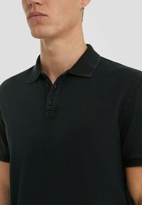 Zwarte polo shirt van een gestructureerde stof, met een knoopsluiting van drie knopen en een kraag. Heeft decoratieve stiksels op de kraag en de knoopsluiting.