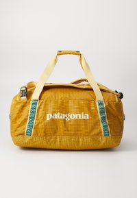 Žuta tekstilna torba na kotačima s tkanim ručkama, Patagonia logo i uzorčani zeleni detalji na bočnom remenu. Glatka tekstura i pravokutni oblik.