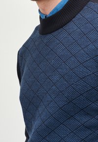 Uomo che indossa un maglione a maglia blu con un motivo sopra una camicia colletto azzurro chiaro, con un girocollo a coste scuro.