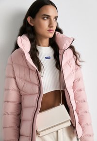 Ροζ φουσκωτό μπουφάν με ψηλό γιακά, με οριζόντιες ραφές. Φορεμένο πάνω από ένα λευκό ριγέ cropped top με μικρό λογότυπο και μια κρέμα τσάντα ώμου.