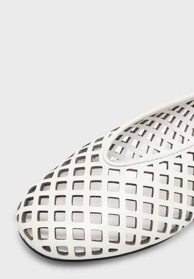 Chaussure blanche sans lacets avec un motif en treillis de découpes carrées et un bout arrondi sur un fond clair uni.