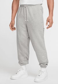 Grå sweatpants med snor i taljen, tætsiddende ankler, sidelommer og et lille logo på venstre ben, parret med hvide sneakers.