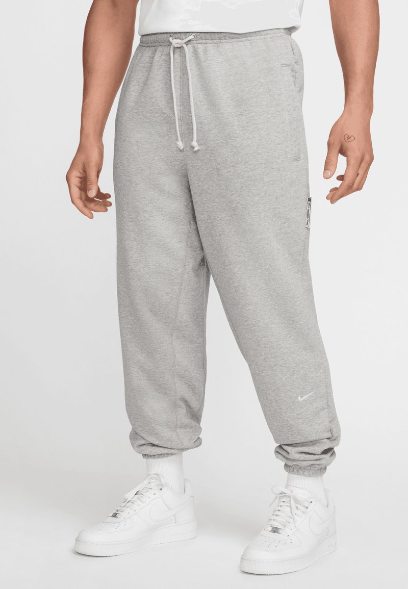 Grå sweatpants med snor i taljen, tætsiddende ankler, sidelommer og et lille logo på venstre ben, parret med hvide sneakers.