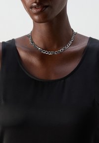 Collar de cadena de metal con picos, de color plateado, llevado sobre piel desnuda; diseño elegante que complementa una blusa sin mangas negra.