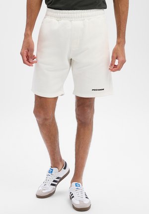 Man draagt witte shorts met elastische taille en het "PEGADOR"-logo, gecombineerd met witte Adidas sneakers met zwarte strepen en een blauw logo.