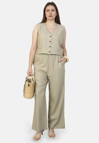 Beige mouwloos topje met knopen, gecombineerd met een broek met wijde pijpen. Model met een geweven handtas. Een gladde textuur; casual ensemble.