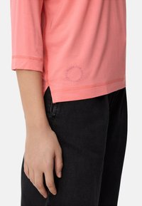 Lyserød top med trekvartærmer, rundet kant og sideslids. Har diskret branding nær kanten. Parret med mørke jeans.