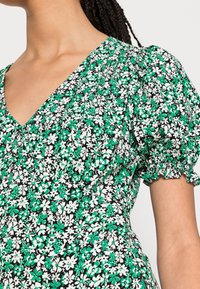 Zeleno-černé květované šaty s nafouknutými rukávy, které mají výstřih do V a nařasené detaily. Materiál má měkký, texturovaný povrch.