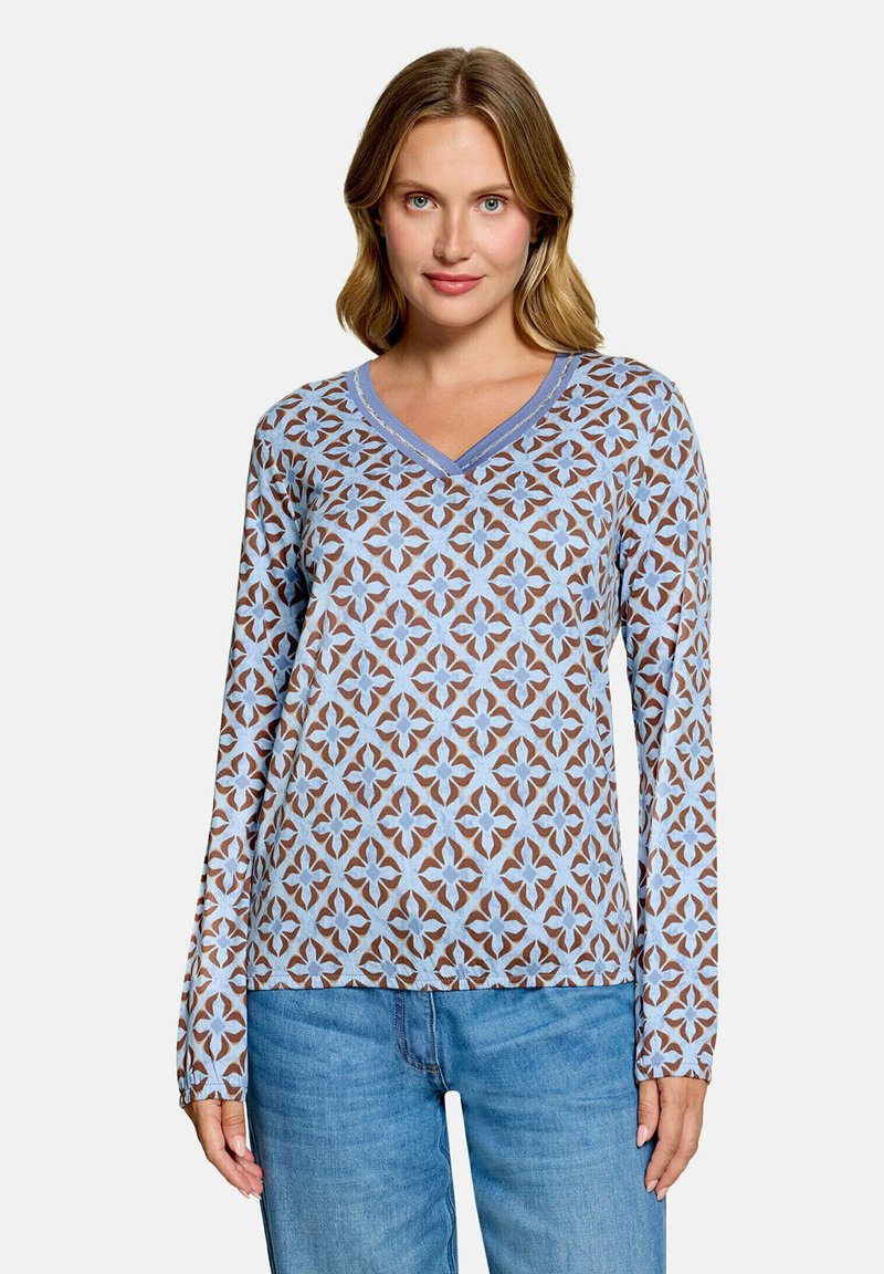 Femme portant un pull manche longue à col en V avec un motif floral géométrique bleu et marron, et un jean bleu, debout devant un fond uni.