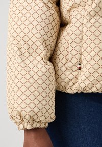 Giacca imbottita di un beige chiaro con un motivo a diamante marrone. Presenta polsini elastici e un'etichetta logo a contrasto sul lato.