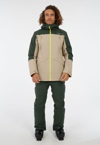 Bežová a zelená lyžiarska bunda s kapucňou, žltým zipsom a dvoma prednými vreckami, skombinovaná so zelenými lyžiarskymi nohavicami a čiernymi lyžiarskymi topánkami.