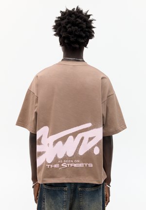 Hnedé oversized tričko s krátkymi rukávmi, s veľkým ružovým textom v graffiti štýle na chrbte. Hladký bavlnený materiál, voľný strih.