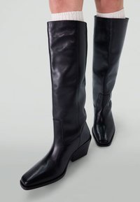 Bottes en cuir noir jusqu'au genou avec un bout carré et un talon carré. Texture lisse et design minimaliste, avec un détail cousu sur le côté.