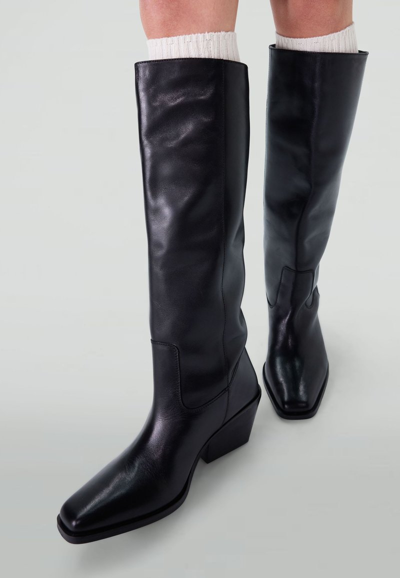 Bottes en cuir noir jusqu'au genou avec un bout carré et un talon carré. Texture lisse et design minimaliste, avec un détail cousu sur le côté.