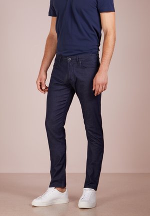 Homme debout portant un jean slim bleu foncé, un t-shirt bleu marine et des baskets blanches sur un fond uni.