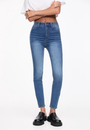 Jean skinny taille haute en denim bleu, avec une légère décoloration, cinq poches et une fermeture classique à bouton et zip.