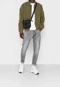 Olivgrön bomberjacka, vit t-shirt, slitna grå jeans och vita sneakers med svarta detaljer. Svart axelremsväska med en logotyp.