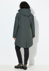 Parka impermeabile di colore verde scuro con cappuccio, maniche lunghe e apertura posteriore per la ventilazione, abbinata a jeans neri e stivali chunky.