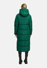 Person with blonde hair in a low ponytail wearing a long, quilted green hooded coat, black pants, and black ankle boots, standing facing away.