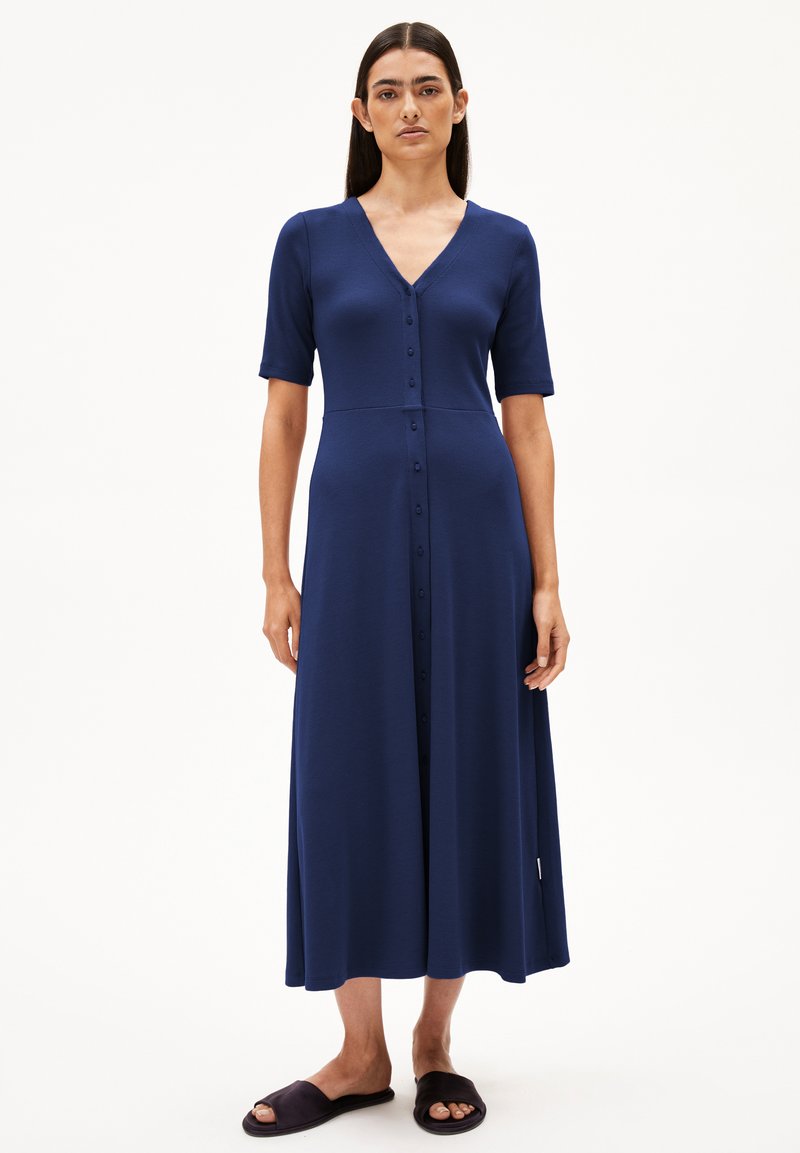 Robe bleu marine, arrivée au genou, à manches courtes, décolleté en V, fermeture à boutons sur le devant, texture lisse et jupe fluide. Associée à des chaussures sombres à enfiler.