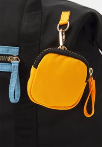 Černý batoh s modrými zipovými detaily a odnímatelnou, čtvercovou, žlutou kapsou, která je připevněna zlatou sponou a oranžovým popruhem.
