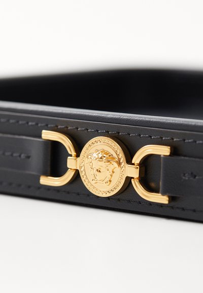 Bolso de cuero negro con herrajes dorados, que presenta un medallón circular y un intrincado estilo de costura en los lados.