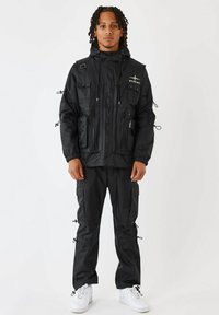 Svart vattentät jacka med flera fickor och dragkedjor, matchad med byxor med fickor. Har dragskor vid muddar och nederkant. Vita sneakers.