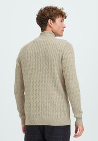 Maglione beige lavorato a maglia con colletto alto, caratterizzato da un motivo a trecce testurizzato e polsini e orlo a costine. La vista posteriore mostra un design aderente e un tessuto morbido.
