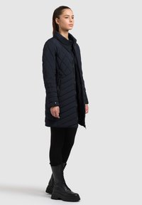 Kvinde iført en marineblå quiltet jakke med lynlås, sorte leggings og sorte snørestøvler, stående mod en ensfarvet lys grå baggrund.