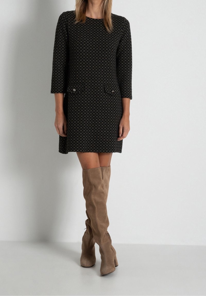 Femme portant une robe noire à motifs avec des manches trois-quarts et des bottes en daim beige montant jusqu'aux genoux, debout contre un mur blanc uni.