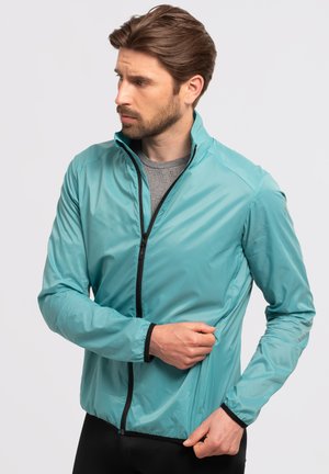 Une veste en bleu sarcelle clair, fabriquée dans un matériau lisse et brillant, présente un col haut, une fermeture éclair à l'avant et des accents noirs au niveau des poignets et de l'ourlet.