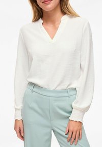 Femme portant un chemisier blanc texturé à manches longues avec des poignets froncés et un pantalon taille haute bleu clair, debout devant un fond uni.