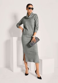 Frau in grauem Strickpullover und passendem Midrock mit Schlitz, schwarzen High Heels, Sonnenbrille, Kreolen und einer gemusterten Clutch.