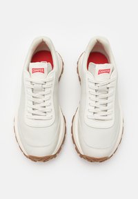 Bílé tenisky s hladkým materiálem, gumovou podrážkou a hnědým podešvem. Mají tkaničky a červené logo Camper na jazyku.