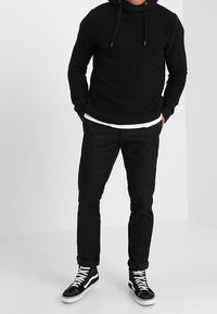 Felpa nera testurizzata con collo alto e cordini, abbinata a pantaloni neri e sneakers alte nere con accenti bianchi.