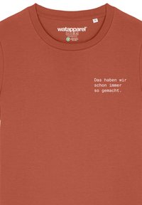 T-shirt en coton couleur rouille avec un col rond. Présente un texte blanc sur le côté gauche : "Das haben wir schon immer so gemacht."