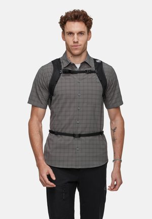 Homme portant une chemise à manches courtes grise à carreaux et un sac à dos noir avec des sangles de poitrine et de taille, debout devant un fond blanc.