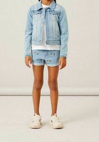 Ljusblå denimjacka med knapprad och fickor, kombinerad med slitna denimshorts. Vita sneakers med texturerad sula.