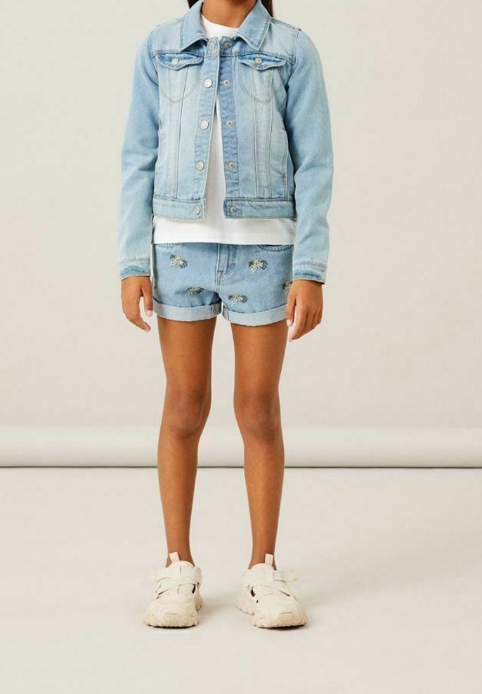 Ljusblå denimjacka med knapprad och fickor, kombinerad med slitna denimshorts. Vita sneakers med texturerad sula.