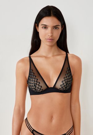 Vrouw met lang zwart haar, gekleed in een zwarte mesh en geborduurd lingerieset, staande tegen een effen lichte achtergrond.